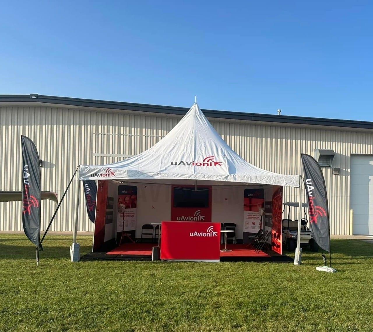 A white promotional tent with red accents, set up outdoors on grass under a clear blue sky.